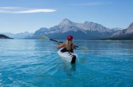 blonde woman kayaking on clear water with mountains in the background